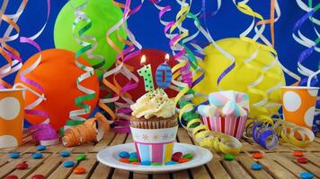 Birthday 10 cupcake with candles burning on rustic wooden table with background of colorful balloons, plastic cups and candies with blue wall in the background photo