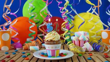 Birthday 52 cupcake with candles burning on rustic wooden table with background of colorful balloons, plastic cups and candies with blue wall in the background photo