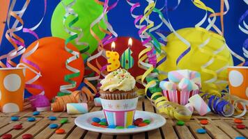 Birthday 36 cupcake with candles burning on rustic wooden table with background of colorful balloons, plastic cups and candies with blue wall in the background photo