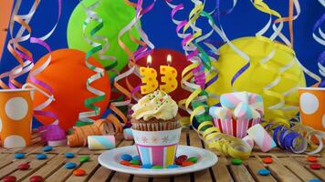 Birthday 33 cupcake with candles burning on rustic wooden table with background of colorful balloons, plastic cups and candies with blue wall in the background photo