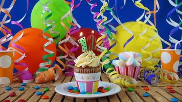 Birthday 12 cupcake with candles burning on rustic wooden table with background of colorful balloons, plastic cups and candies with blue wall in the background photo