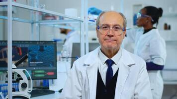 Portrait of senior experienced scientist man smiling at camera in modern equipped lab. Multiethnic team examining virus evolution using high tech and chemistry tools for scientific research, vaccine development. photo