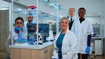 Team of multiethnic scientists sitting in laboratory looking at camera in modern equipped lab. Group of doctors examining virus evolution using high tech for scientific research, vaccine development. photo