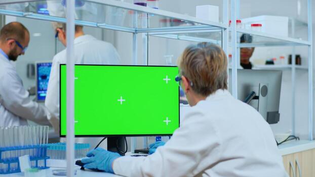 Elderly researcher looking at chroma key display in modern equipped lab. Multiethnic team of microbiologists doing vaccine research writing on device with green screen, isolated, mockup display. photo