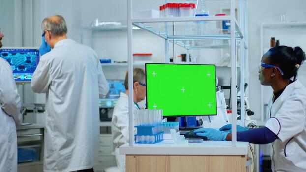 African scientist woman typing on computer with green mockup in modern equipped lab. Multiethnic team of microbiologists doing vaccine research writing on device with chroma key, isolated display. photo