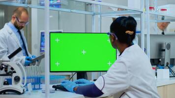 African doctor working at computer with green screen in modern equipped lab. Multiethnic team of microbiologists doing vaccine research writing on device with chroma key, isolated, mockup display. photo