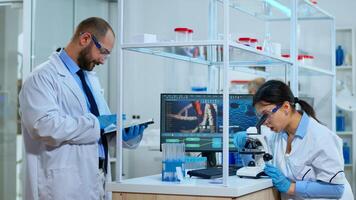 Team of scientists doing biological researches under microscope, lab technician writing data into tablet in modern equipped laboratory. Multiethnic group examining virus evolution using high tech. photo