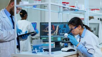 Scientist team checking virus development using microscope and writing analysis results on tablet in modern equipped lab. Multiethnic group examining vaccine evolution using high tech for research. photo