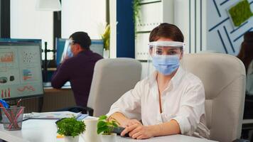 Businesswoman with visor and mask talking in front of camera having call with partners. Pov of manager speaking with remotely team during online conference while colleagues working in background photo