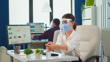 Entrepreneur with protection mask and visor applying sanitizer gel rubbing hands before typing at computer. Businesswoman in new normal workplace disinfecting while colleagues working in background photo