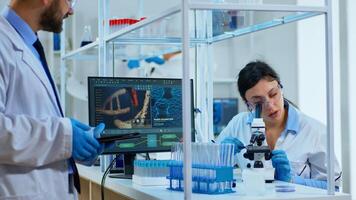Team medical scientist conducting DNA experiments under digital microscope writing results into tablet working in science laboratory. Caucasian lab engineer in white coat analysing vaccine development photo