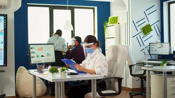 Teamworkers in new normal office making financial strategy wearing protective mask. Employees with visors working in corporate company workspace respecting social distance analysing data and graphs. photo