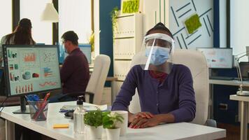 African businesswoman with visor and mask talking at camera having meeting with remote partners. Pov of woman speaking with team during online conference while colleagues working in background photo
