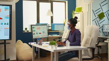 Black employee with protection mask and visor cleaning hands with sanitizer gel before writing at computer. Businesswoman in new normal workplace disinfecting while colleagues working in background photo