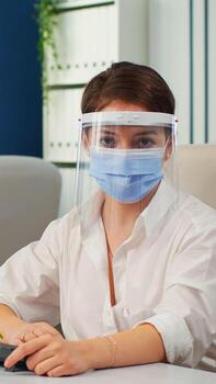 Businesswoman with visor and mask talking in front of camera having call with partners. Pov of manager speaking with remotely team during online conference while colleagues working in background photo
