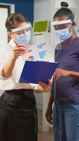 Company manager with visor against coronavirus explaining to african colleague project plan standing in front of desk holding clipboard. Multiethnic business team working respecting social distance. photo
