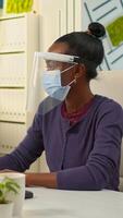 African company manager with face mask writing on computer and looking on camera in new normal business financial office respecting social distance. Multiethnic colleagues working in background. photo