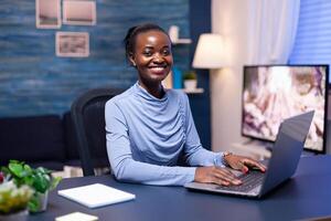 Smiling dark skinned woman smiling at camera sitting at desk working late at night from home office. Black freelancer working with remotely team chatting virtual online conference. photo