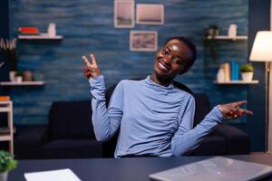 Cheerful dark skinned business woman showing victory sign looking at camera working late at night from home. . Black woman enjoy e-learning process, easy application usage overtime. photo