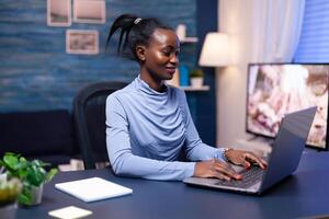 Dark skinned business woman working hard to finish a deadline using laptop computer from home late at night. Black entrepreneur sitting in personal workplace writing on keyboard. photo