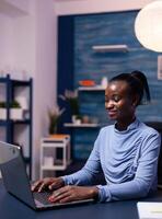 African business woman working on laptop trying to finish a deadline for work from home office late at night. Black entrepreneur sitting in personal workplace writing on keyboard. photo