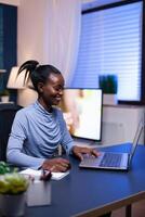Dark skinned freelancer in the course of webinar smiling while taking notes sitting at desk late at night. Black entrepreneur sitting in personal workplace writing on keyboard. photo