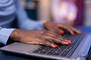 Close up of dark skinned business woman hand typing email n laptop computer from home office late at night. Employee using modern technology network wireless doing overtime. photo