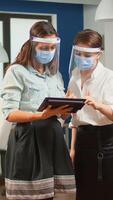 Businesswomen with protection masks against coronavirus standing in workplace talking about company financial datas holding digital tablet. Multiethnic business team working respecting social distance photo