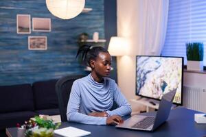 Dark skinned freelancer reading documentation on laptop computer in living room late at night. Black entrepreneur sitting in personal workplace writing on keyboard. photo