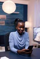 Cheerful african business woman texting on smartphone late at night in home office working remote. Employee using modern technology network wireless doing overtime. photo