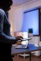 Dark skinned woman using tablet pc keybpad to write email working from home office. Busy focused employee using modern technology network wireless doing overtime writing. photo