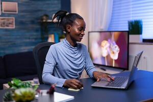 Dark skinned woman taking notes while working on deadline using laptop sitting at desk from home in the evening. Black entrepreneur sitting in personal workplace writing on keyboard. photo