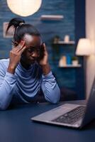 Overworked african business woman having a headache while working late in the night from home office. Tired focused employee using modern technology network wireless doing overtime. photo