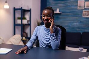 African business woman speaking on smartphone working late at night from home office. Busy focused freelancer using modern technology network wireless doing overtime. photo