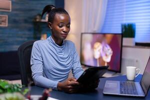 Afro woman working remote from home office late in the evening using tablet pc. Busy focused employee using modern technology network wireless doing overtime writing. photo