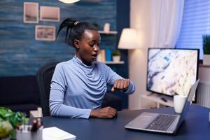 African business woman checking time looking at wrist watch working late on deadline from home office. Nervous black manager doing overtime working on computer. photo