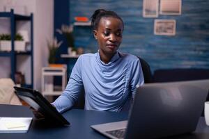Concentrated african woman working on deadline using tablet pc and laptop in home office late at night. Busy focused employee using modern technology network wireless doing overtime writing. photo