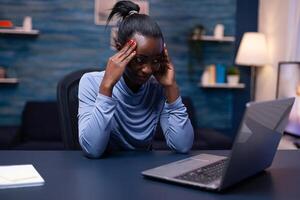 Dark skinned woman touching foarhead because of headache while working from home. Tired focused employee using modern technology network wireless doing overtime. photo