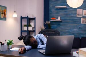 African business woman sleeps during late night hourse in the course of deadline working from home. Busy employee using modern technology network wireless doing overtime sleeping on table. photo