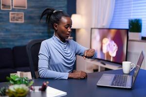 Worried african entrepreneur looking at wrist watch working hard to finish a big project for work. Nervous black manager doing overtime working on computer. photo