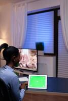 African entrepreneur sitting at desk in home office holding tablet pc with green screen working on deadline. Using mockup chroma key display computer. photo