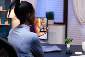 Dark skinned woman talking about project with coworkers late at night in the course of conference on smartphone. Busy employee using modern technology network wireless doing overtime for job. photo