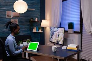African businesswoman looking at tablet pc in the course of working from home late at night in home offce sitting at desk. Using mockup chroma key display computer. photo
