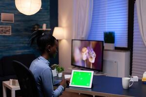 Black entrepreneur woman working on project deadline from home at night in home office holding tablet pc with green screen. Using mockup chroma key display computer. photo