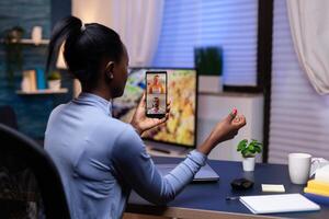 Dark skinned woman using smartphone late night in home office discussing wtih people in conference. Busy employee using modern technology network wireless doing overtime for job. photo
