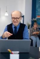 Happy senior man pointing at laptop during conference. Cheerful pensioner with gray hair smiling at laptop webcam while chatting sitting at desk . photo