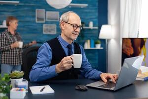 Senior businessman holding cup of coffee working on laptop. Elderly man entrepreneur in home workplace using portable computer sitting at desk while wife is holding tv remote. photo