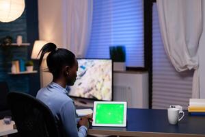 Black freelancer woman working from home late at night with device having copy space available sitting at office.Using mockup chroma key display computer. photo