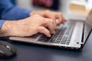 Close up of senior man hands typing on laptop keyboard. Elderly man entrepreneur in home workplace using portable computer sitting at desk while wife is holding tv remote photo