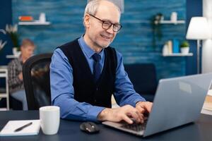 Smart senior businessman working on laptop wearing a tie and glasses. Elderly man entrepreneur in home workplace using portable computer sitting at desk while wife is holding tv remote. photo